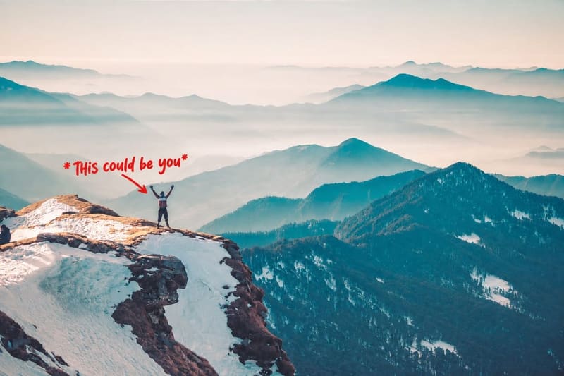 A climber standing with arms raised on a snowy summit above a wide range of misted peaks