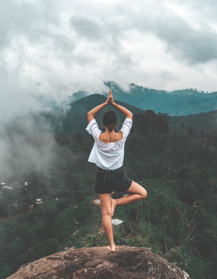 A woman balancing in a yoga pose on a rock above a wooded valley